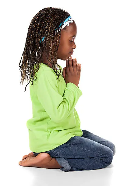 Adorable small african child with braids wearing a bright green shirt and blue jeans. The girl is kneeling and praying.