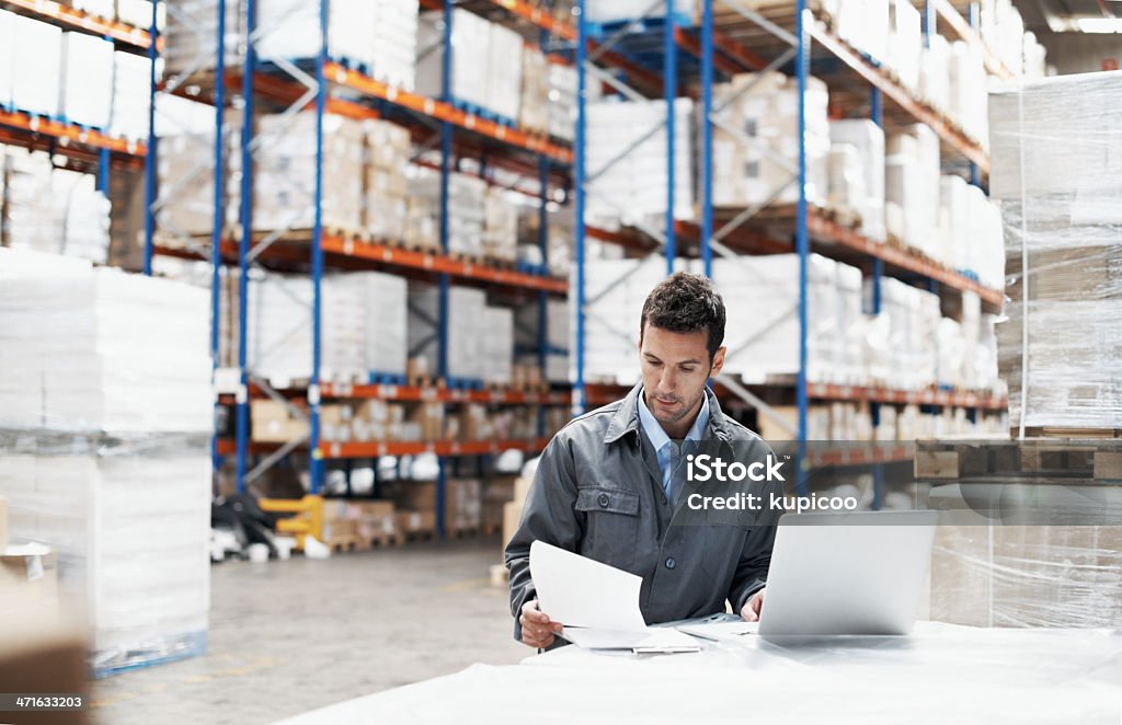 Prioritising the deliveries A young man using his laptop and looking over his notes while working in a warehouse Warehouse Stock Photo