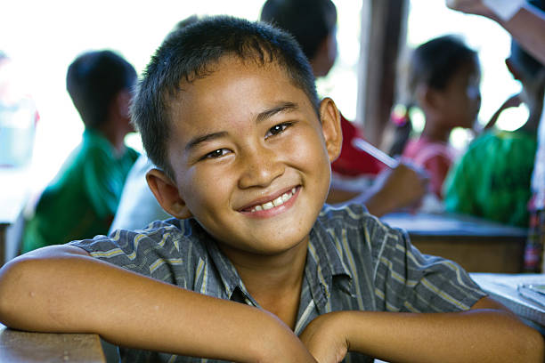 A young Asian boy sits in a classroom and is smiling at the camera.