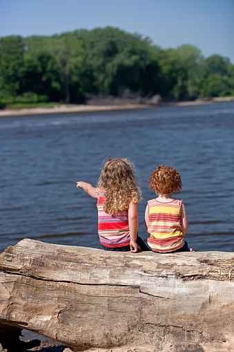 Two young sisters sitting on a large log looking out at the Mississippi River. The older girl is pointing something out to the younger girl.