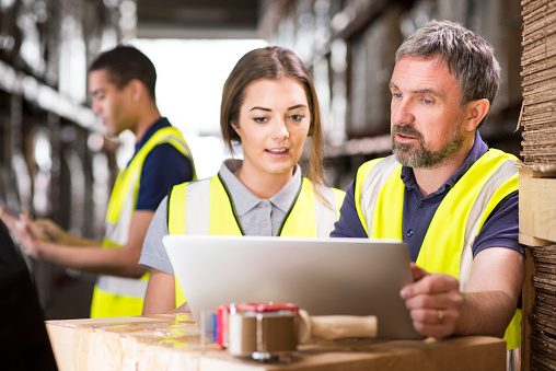 a mature male warehouse manager is chatting to a young female colleague in front of a laptop. behind them can be seen a large warehouse interior , and a male colleague slightly defocussed.