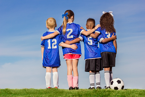 Diverse group of boys and girls soccer players standing together with a ball against a simple blue sky background.