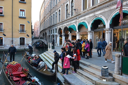 Venice, Italy - March 12, 2015: A group of about 20 asian tourists queuing for rides on Gondolas on the canals of Venice. Other various adults walk by on the right. Gondola operators man the boats. 