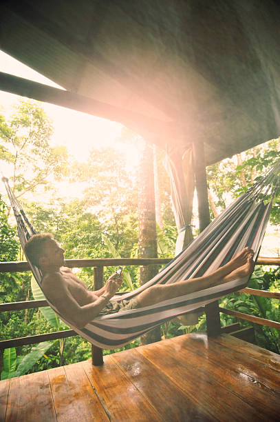 istock Man Relaxes  Sunny Jungle Lodge Hammock with Smartphone Mosquito Net 471649779