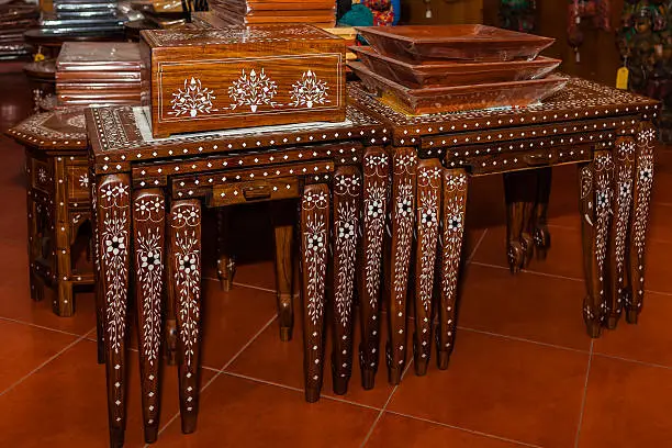 Indian handicraft inlaid rosewood coffee tables that fit into each other to save space, for sale.  The inlay work creates intricate patterns on the carved tables. Tourists to India, buy them as souvenirs that can be actually used in a home. Focus on the table legs in the foreground; shallow depth of field.  Horizontal format. No people.