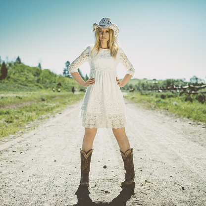 All-American Girl in White Dress, Cowboy Boots and Cowboy Hat standing in the Middle of s dusty Road. Cross-processed, squared lifestyle portrait.