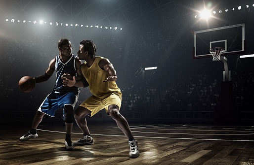 Low angle view of a professional basketball game in 80's. The colors are desaturated. A player failed to block  the opposite team player with a ball. A game is in a indoor floodlit basketball arena. All players are wearing generic unbranded basketball uniform.