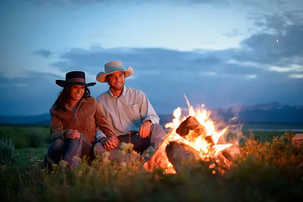Young American cowboy couple enjoy the Western wilderness and the warm glow of a campfire together at dusk.