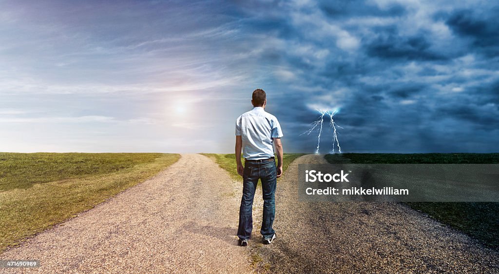 Man must decide his way forward to success or failure Concept of personal decision making. Man stand at a forked road and has to choose his way forward. The one to left is covered in sunlight. The one to the right is dark and lightning is in the horizon. Forked Road Stock Photo
