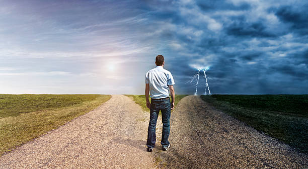 Man must decide his way forward to success or failure Concept of personal decision making. Man stand at a forked road and has to choose his way forward. The one to left is covered in sunlight. The one to the right is dark and lightning is in the horizon. road to success stock pictures, royalty-free photos & images