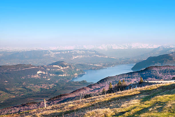 Horizontal scenics landscape view of Bourget lake with Aix les Bains city (in Savoie) in background, and Mont Blanc massif, from top mountain peak, of Alps chain in end of summer, autumn season. Photography taken on Grand Colombier mountain (near Culoz city), in Bugey, in Ain, Rhone-alpes region in France (Europe). Shot with some grass and trees.