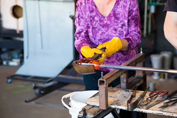 A woman is shaping some motlen glass into a bowl using a wooden forming tool at a glass studio.