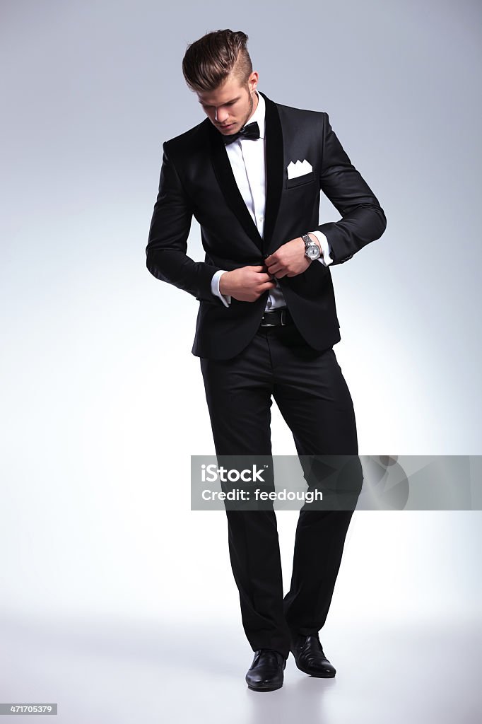 business man buttoning and looking down full length picture of an elegant young fashion man in tuxedo buttoning his jacket while looking down, away from the camera. on gray background Adult Stock Photo