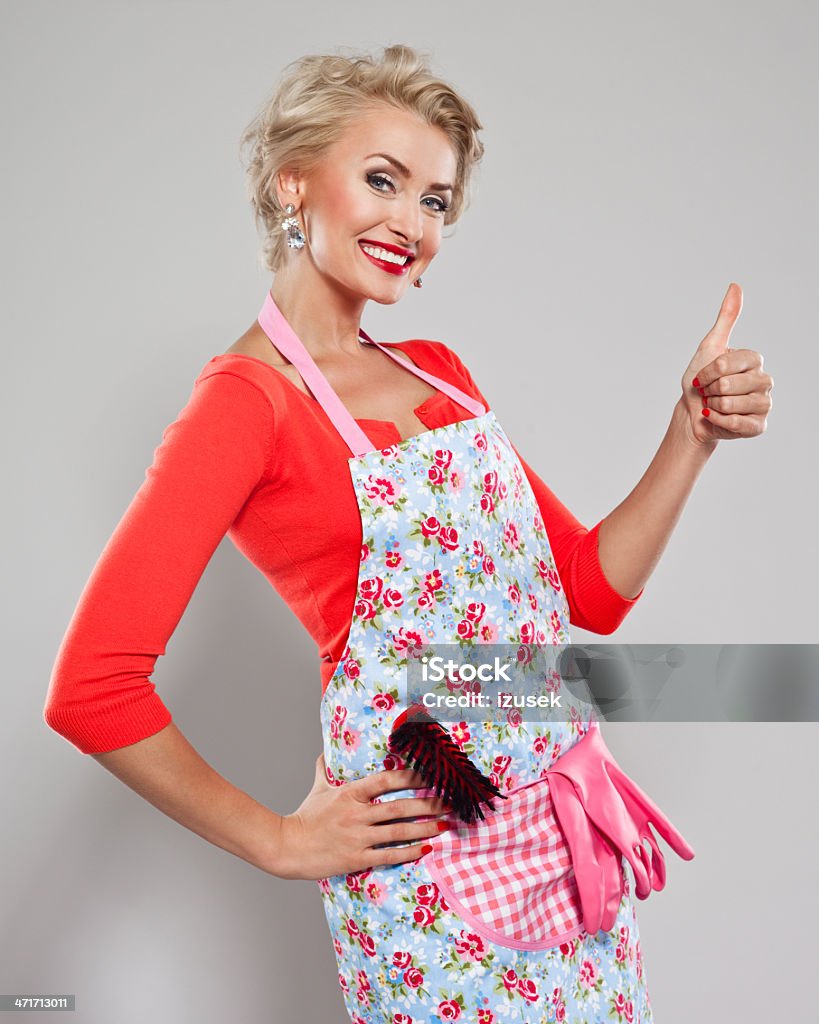 Elegant cleaner Portrait of a cheerful housewife wearing an apron with broom and washing up gloves in pocket and smiling at the camera with thumb up. 30-34 Years Stock Photo