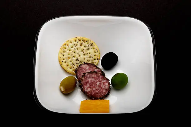 selection of cheddar cheese,peppered hard salami, a sesame cracker and three colorful olives in a white bowl