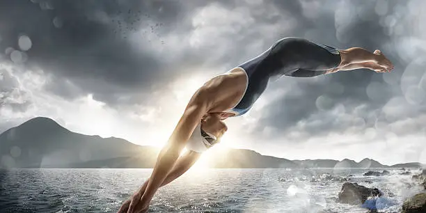 A mid action image of a professional female swimmer dressed in generic swimming bodysuit, swim hat, and goggles in mid air diving into the sea close to some rocks. The swimmer is in a generic location near mountains under a stormy evening sky. With intentional water splash bokeh and lensflare.