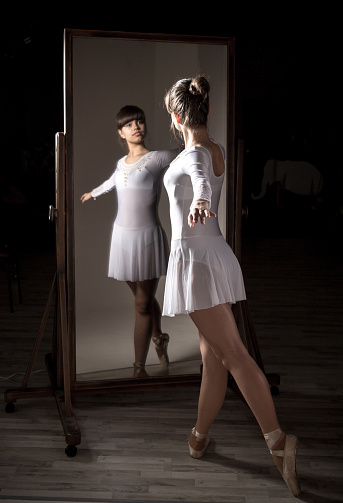 Beautiful girl ballet dancer standing before a full-length mirror in a dark studio. Young ballerina looking to her reflection in the mirror