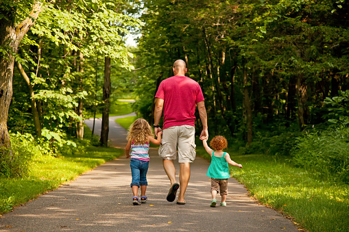 Father and his two young daughters walking on a paved path through the park. They are all walking side by side and holding hands with each other.
