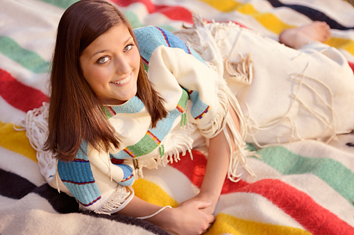 Beautiful Indian Maiden in authentic deer skin, leather beaded, traditional dress.  She is laying on a  multi-colored authentic, hand woven blanket.  She is smiling and looking up at the camera.  Barefoot.