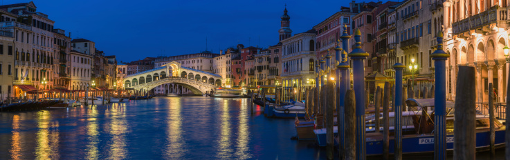 Locals and tourists on the iconic stone arch of Rialto Bridge illuminated in the blue dusk above the broad sweep of the Grand Canal between the busy restaurants, historic palazzo, gondolas and boats of the San Marco and San Polo quarters of Venice, Italy.  ProPhoto RGB profile for maximum color fidelity and gamut.