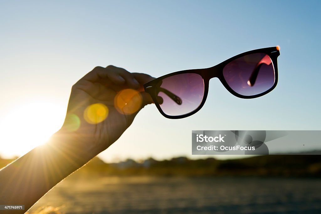 Backlit Hand and Sunglasses 02 Hand Holding Black Sunglasses at sunset in front of vibrant blue sky. The Sun iluminates de scene with the golden light of dawning.  Backlit UV Protection Stock Photo