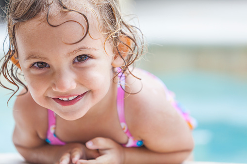 Portrait of multiracial elementary-age girl with curly hair wearing swim goggles swimming in a pool on a sunny day.