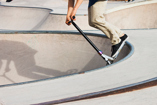 Boy in a concrete skate park with a push scooter making a trick in the rail of a bowl.