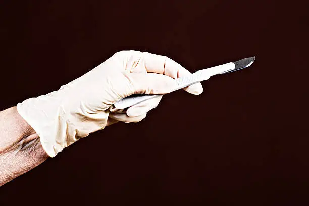 The gloved hands of an elderly medical professional hold up a surgical scalpel ready to slice into something. Dark background with copy space.