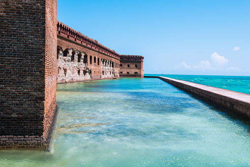 This is a horizontal, color toned photograph of the historic brick fortress, Fort Jefferson on Garden Key in the Dry Tortugas National Park in Florida. A wall surrounds the former military outpost. Tropical colored salt water fills the moat and surrounding wall. 