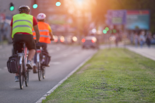 Two cyclists with protective equipment are approaching an intersection in a busy part of the city towards the setting sun. Shallow depth of field, focus on the grass in the foreground.
