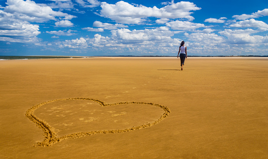 A young woman walks away from a love heart drawn in the sand on a beach