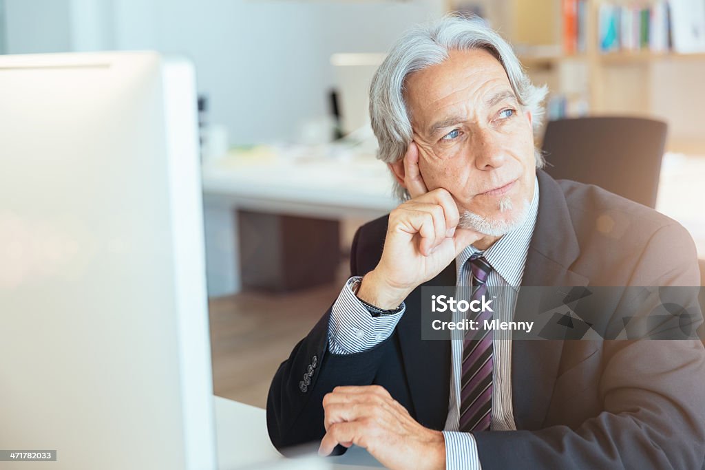 Senior Businessman Thinking Senior businessman sitting at his work desk behind a large flat screen, looking thoughtfully through the window into the sunlight. Contemplation Stock Photo