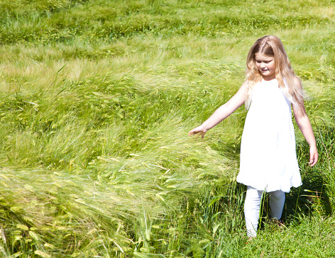 Seen in a panoramic crop, a little blonde girl plays outdoors on grass, looking down and absorbed in something.