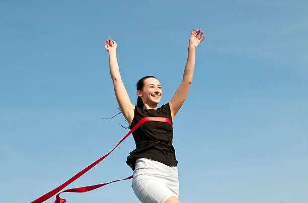sucessful young business woman crossing red finish line ribbon, against blue sky