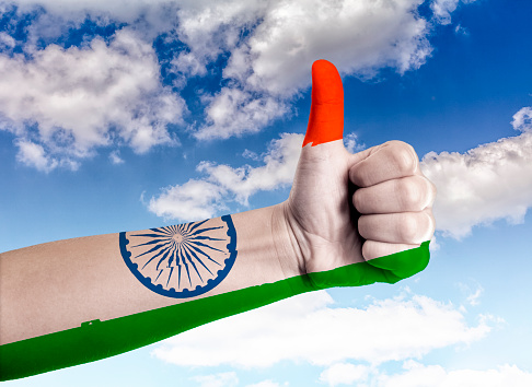 Hand, holding up a thumb with indian flag painted on it, against blue sky with cloudscape.