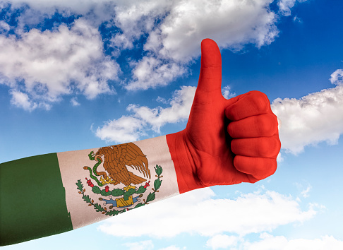 Hand, holding up a thumb with mixican flag painted on it, against blue sky with cloudscape.