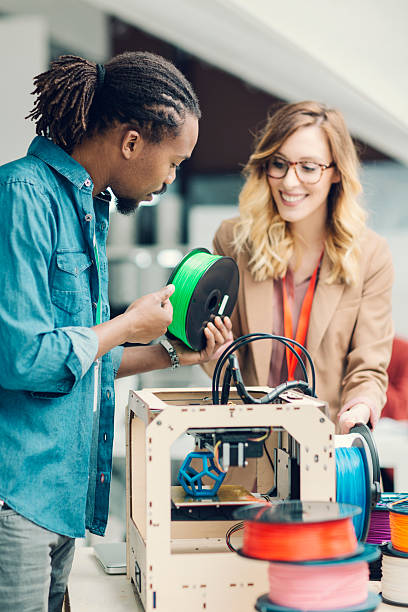 Mixed Race Woman and Man standing by 3D Printer. Diversity startup business team working together by 3D printer. Looking at camera and holding 3D printer filaments. He is holding green filament and she is holding yellow filament. They are standing by 3d printer. 3d printing photos stock pictures, royalty-free photos & images