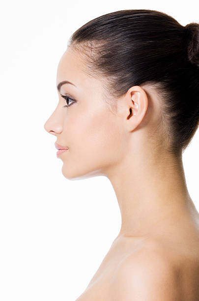 A woman is standing in front of the camera, facing the left side of the image, isolated against a white background.  She has dark brown hair that is pulled back high at the back of her head into a bun.  Her shoulders and upper chest are bare.