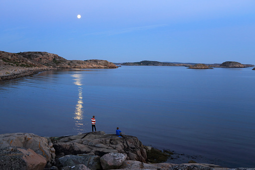 Two boys night fishing on archipelago island under a full moon. Still almost light outside due to the late sunset during summer in the Nordics.