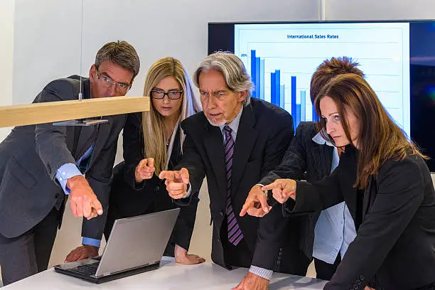 Mixed group of business people finger pointing at one (invisible) colleague in a meeting with current sales figures projected on a screen in the background