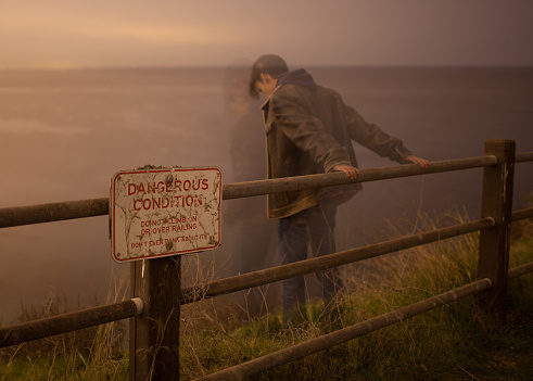 Depressed man looking over edge of cliff, danger sign on left. Taken in low light with long exposure and low ISO.