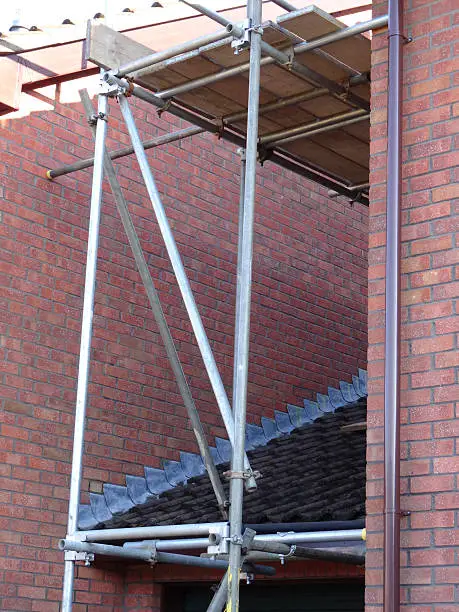 Photo showing a modern, red-brick house that is having some repairs and construction work carried out to its roof, with builders using a small scaffolding tower to safely carry out their work from.