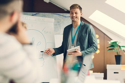 Young smiling man has briefing with his coworkers at startup creative agency.He is standing in front of flip chart, holding digital tablet and talking about creativity. Looking at coworkers and smiling.