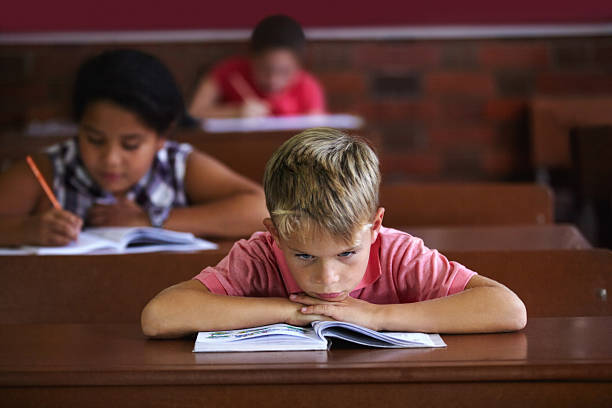 School sucks! A young boy resting his head on his arms as he sits in a classroom looking bored gloomy mood stock pictures, royalty-free photos & images