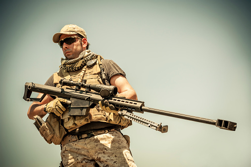 Contemporary marksman is walking with .50 caliber precision rifle.