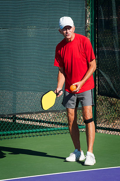 Pickleball Action - Senior Man Preparing To Serve Senior man in bright red shirt preparing to serve pickleball.  Announcing the score. knee brace stock pictures, royalty-free photos & images