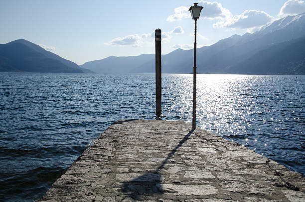 Pier on an alpine lake with mountains and street lamp with shadow in ticino switzerland