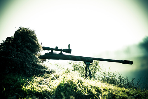 A marksman on grass, dressed with a camouflage ghillie suit, is taking aim with his scope mounted on a precision rifle before shooting.