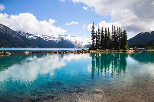 The clear, tranquil waters of Garibaldi Lake in Garibaldi Provincial Park near Whistler in British Columbia, produce bottle-green reflections of an island of conifers.  In the foreground close to shore, small rocks are visible beneath the water surface. As the glacially-fed water gets deeper, it takes on shades of turquoise and emerald.