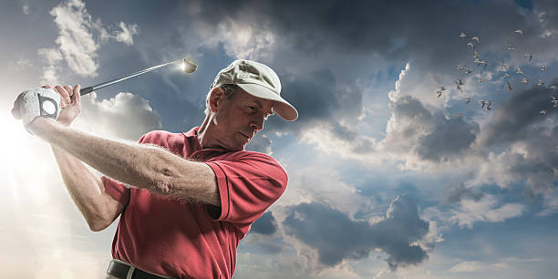 Golfing In The Sun A close up low angle portrait of a senior male golfer from waist up holding a driver club in a backswing ready to strike ball. The golfer is standing against a cloudy blue sky with a low bright evening sun causing lensflare. The golfer is wearing a golfing cap, golfing glove, and generic red polo shirt. blue and white tees stock pictures, royalty-free photos & images